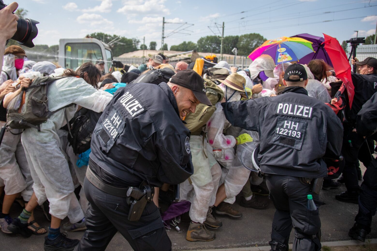 Eine Menschenmenge mit vielen Personen im weißen Overall wird von Polizist*innen mit Pfefferspray angegriffen, ein Beamter bekommt dies ins Gesicht.